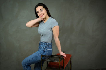 Obraz na płótnie Canvas Portrait of a pretty young brunette woman in a gray t-shirt and blue jeans sits on a stand. The concept of a cute girl on a gray background. Smiling, showing hands with emotions.
