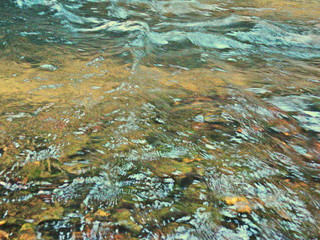 Ripples in the river flow through the rocks, colorful in the summer in bright sunlight, have background of the shiny pebbles, at the bottom of river In a abstract, form of pebbles below water surface.