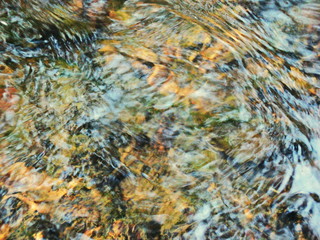 Ripples in the river flow through the rocks, colorful in the summer in bright sunlight, have background of the shiny pebbles, at the bottom of river In a abstract, form of pebbles below water surface.