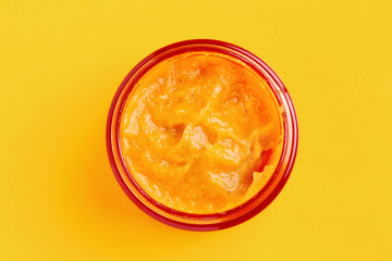 An open glass jar with an orange facial scrub is on a yellow background