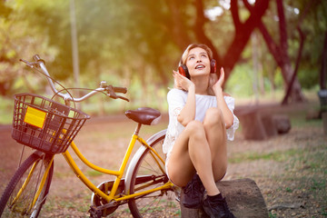 Young cheerful caucasian woman sitting on tree stump with yellow bicycle beside and listening online music from headset with casual style in park during relaxation with happiness face and smile.