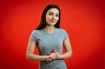 Fototapeta na wymiar Universal concept of a cute smiling girl on a red background. Portrait of a pretty young brunette woman in a gray t-shirt. Talking, showing hands with emotions.