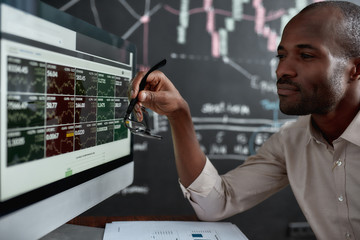 Be the Financial Fit. African businessman, trader sitting in front of computer screen and holding glasses while looking at graph chart. Blackboard full of data analyses in the background. - obrazy, fototapety, plakaty