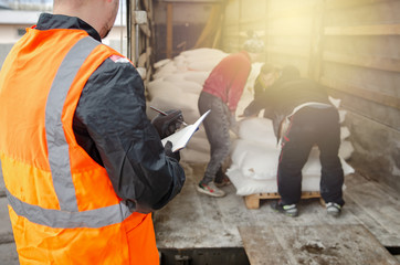 a manager in a protective helmet and overalls controls the unloading and records the number of bags of flour. warehouse logistics cargo courier, shipment transportation, worker holding clipboard check