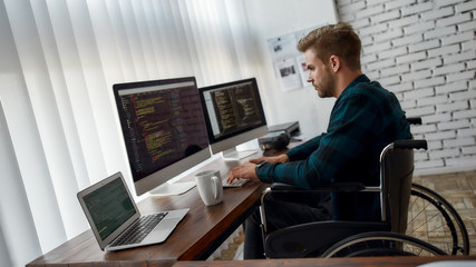 Writing computer program. Side view of young professional web developer in a wheelchair writing program code on multiple computer screens while sitting at his workplace in the modern office