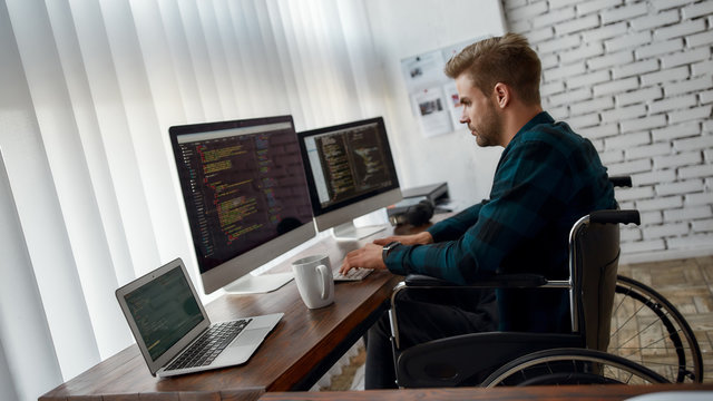 Writing computer program. Side view of young professional web developer in a wheelchair writing program code on multiple computer screens while sitting at his workplace in the modern office