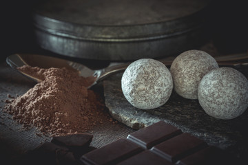 Chocolate truffles covered with powdered sugar nicely arranged on wooden table with crushed dark chocolate bar and cacao power on a spoon. Dark mood photography concept.