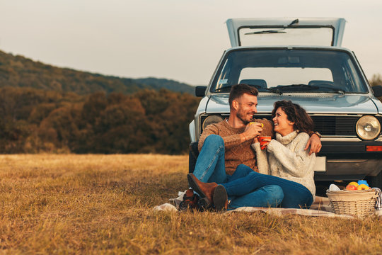 Beautiful young couple enjoying picnic time on the sunset. They drinking tea and sitting in a meadow leaning on old fashioned car.