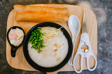 Top view of congee pork in black bowl with onsen egg, sliced spring onion, ginger, deep-fried dough stick and scissors for cutting on wooden tray to serve a breakfast to customer in restaurant