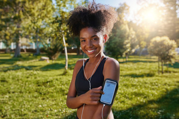 Portrait of a smiling african american young woman touching her mobile phone enjoying listening to music with earphones in the park
