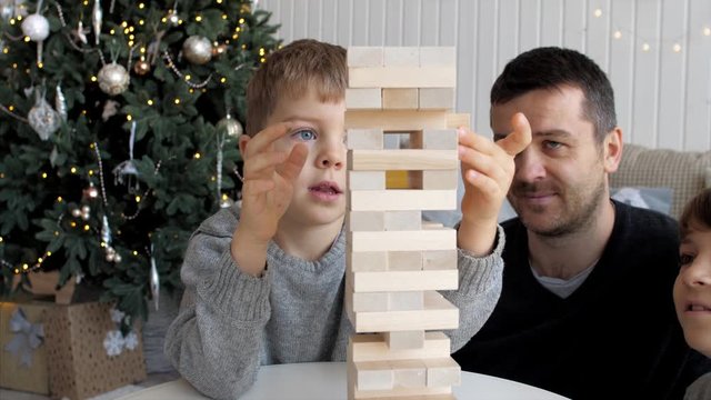 Family is playing in board game with wooden tower together at home. Junior son is pulls out a brick while older brother and father is looking on it.