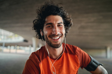Portrait of happy young male runner with earphones in his ears looking at camera outdoor smiling -...