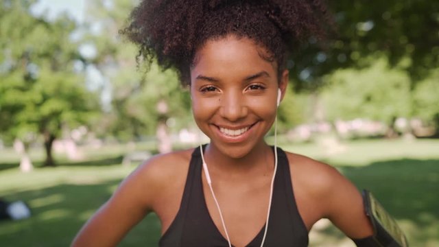 Portrait of a cheerful fit african american young woman listening to music on earphones smiling and looking at camera in the park