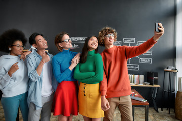 Having fun together. Positive group of multicultural people in casual wear making selfie on the phone while standing in the modern office. Young cheerful designers looking into the camera