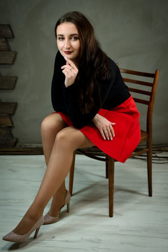 Model in a dark blouse and a red skirt. Vertical Photo of a pretty slender brunette young woman with good make-up sits on a chair in a vintage interior. Smiling happy.