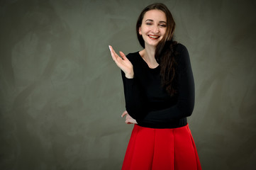 Model in a dark blouse and a red skirt. Photo of a pretty slender young brunette woman with good makeup on a gray background. Talking and showing hands with a smile