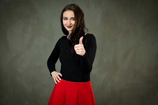 Photo of a pretty slender young brunette woman with good makeup on a gray background. Model in a dark blouse and a red skirt. Talking and showing hands with a smile