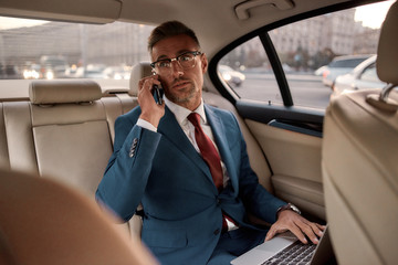 Being good in business. Confident and stylish mature businessman in full suit working on his laptop and talking on the phone with business partner while sitting in the car