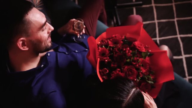 Beautiful young couple in love enjoying each other, olding flowers and champagne on a couch. Smiling in a romantic atmosphere with candles.