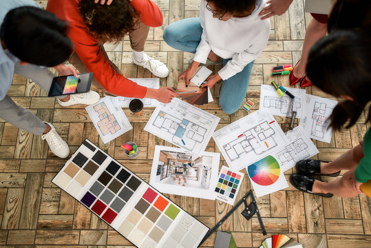 Discussing sketches. Group of young interior designers in casual clothes working on a new project, choosing colors from palettes lying on the floor in the modern office. Creative agency. Design studio - Powered by Adobe