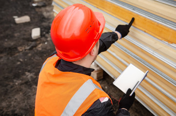  A worker in a protective helmet and overalls makes an inventory of sandwich panels and writes...