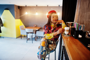 Enthusiastic african american woman in trendy coloured outfit with red beret chilling in cozy cafe, sitting on bar counter.