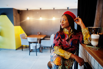 Enthusiastic african american woman in trendy coloured outfit with red beret chilling in cozy cafe, sitting on bar counter.