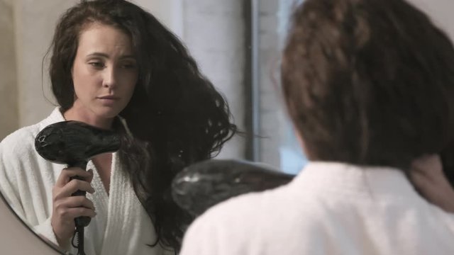 Beautiful Caucasian woman in white bathrobe looking in bathroom mirror and drying her long brunette hair with blow dryer