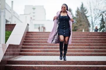 Young stylish beautiful african american woman in street against stairs, wearing fashion outfit coat, eyeaglasses.