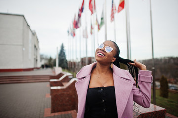 Young stylish beautiful african american woman in street, wearing fashion outfit coat and eyeglasses, against flags of different countries of the world.