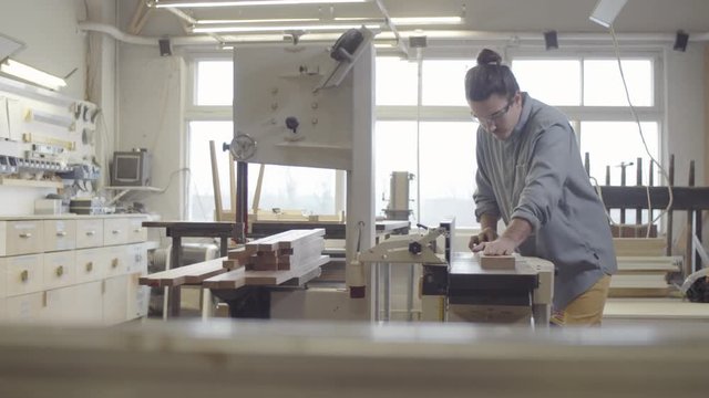 Full shot of young Caucasian male carpenter in safety goggles working at his furniture making workshop and carefully smoothing edges of wood board on jointer machine