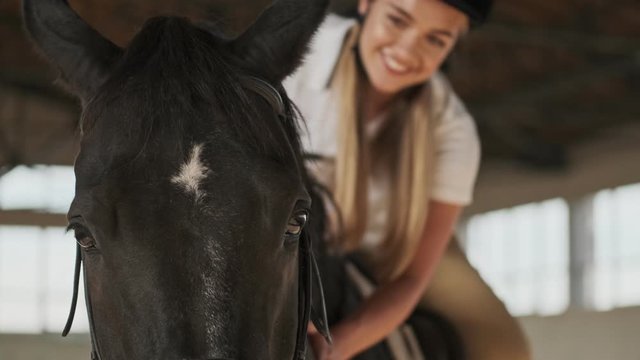 Close up view of a smiling girl is petting a horse while riding it in the covered stable