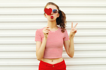 Beautiful young woman with red heart shaped sunglasses blowing lips sending sweet air kiss on white wall background