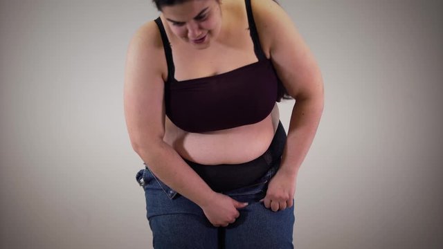 Portrait of positive fat Caucasian girl putting in jeans. Obese young woman jumping trying to pull up trousers. Cellulite, obesity, fatness.