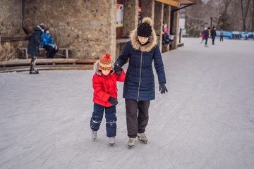 young mother teaching her little son ice skating at outdoor skating rink. Family enjoy winter on ice-rink outdoors