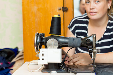 Pretty female tailor sits at table with sewing machine and sews fabric.