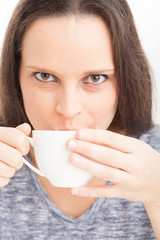 Female drinking cup of warm tea of coffee, wearing blue on white background, looking at camera