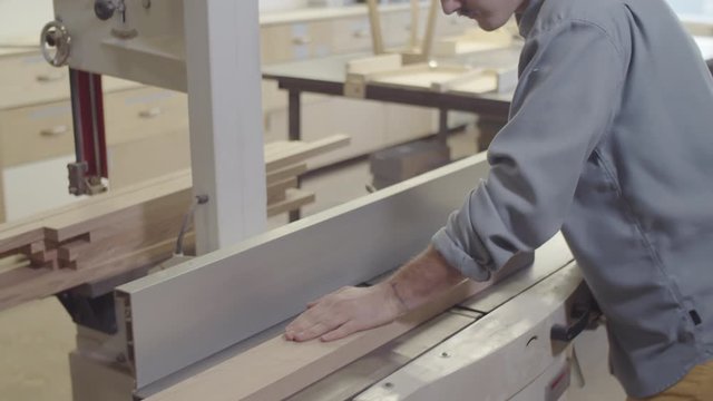 High angle tilting down shot of young man with long hair in bun, in safety goggles smoothing rough surface of wood board on jointer lathe at carpentry workshop