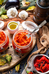Tabletop scene with raw red chutney with a silver knife surrounded with vegetables. Raw Ajvar in a small glass jar