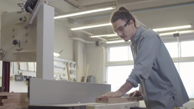 Thigh-up shot of young Caucasian carpenter in safety goggles checking surface of wood board then smoothing rough edge on jointer machine, while working at furniture making workshop
