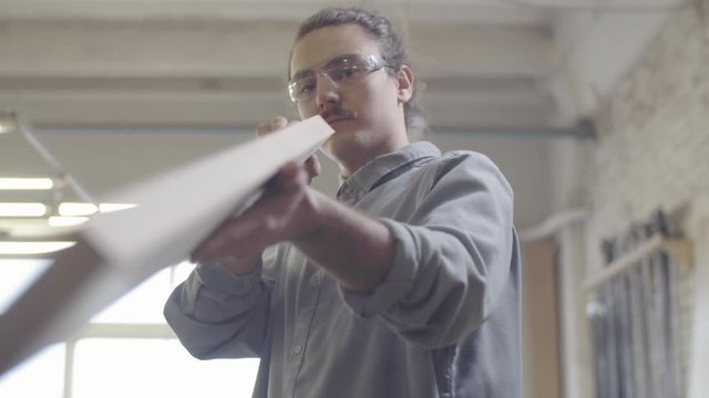Waist-up shot of young woodworking artisan lifting trimmed natural wood board from jointer lathe, lifting it up, checking smooth surface while squinting, and feeling it by hand