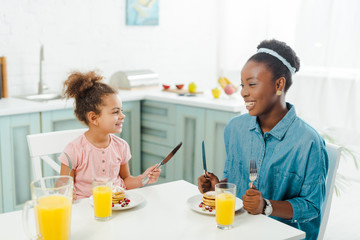 cheerful african american mother and daughter holding cutlery near tasty pancakes