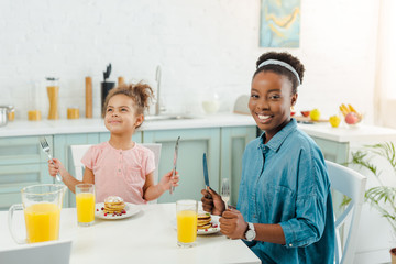 happy african american mother and daughter holding cutlery near tasty pancakes