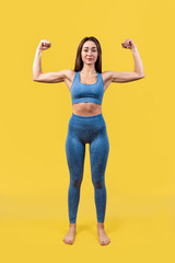 Portrait of beautiful athletic Caucasian woman in blue sportswear showing biceps of her hands. Yellow background. Copy space. Vertical