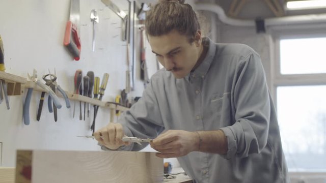 Waist-up shot of young Caucasian man with long hair in bun working in carpentry workshop and carefully measuring and checking thickness of smooth natural wood board with calipers
