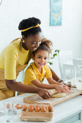 attractive african american mother looking at raw dough near cute daughter and holding rolling pin