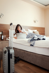 Young businesswoman working from hotel room on business trip, woman lying on the bed and using...