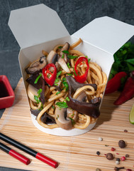 Box for wok. With egg noodles, mushrooms, chili peppers and soy sauce. The wok box is cut. Standing on a mat next to chopsticks, spices and soy sauce. Close-up. Gray background.