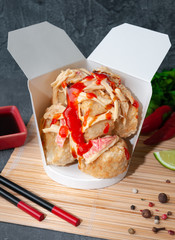 Box for wok. With pieces of fried tofu, crab meat, squid. And hot chili sauce. The wok box is cut. Standing on a mat next to chopsticks, spices and soy sauce. Close-up. Gray background.