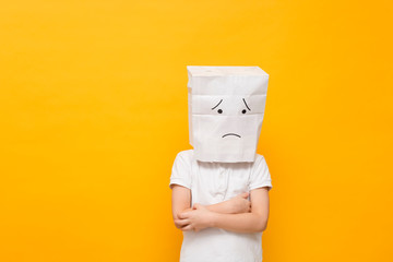 Cute little school boy standing with a paper bag on his head - sad face on yellow background, concept